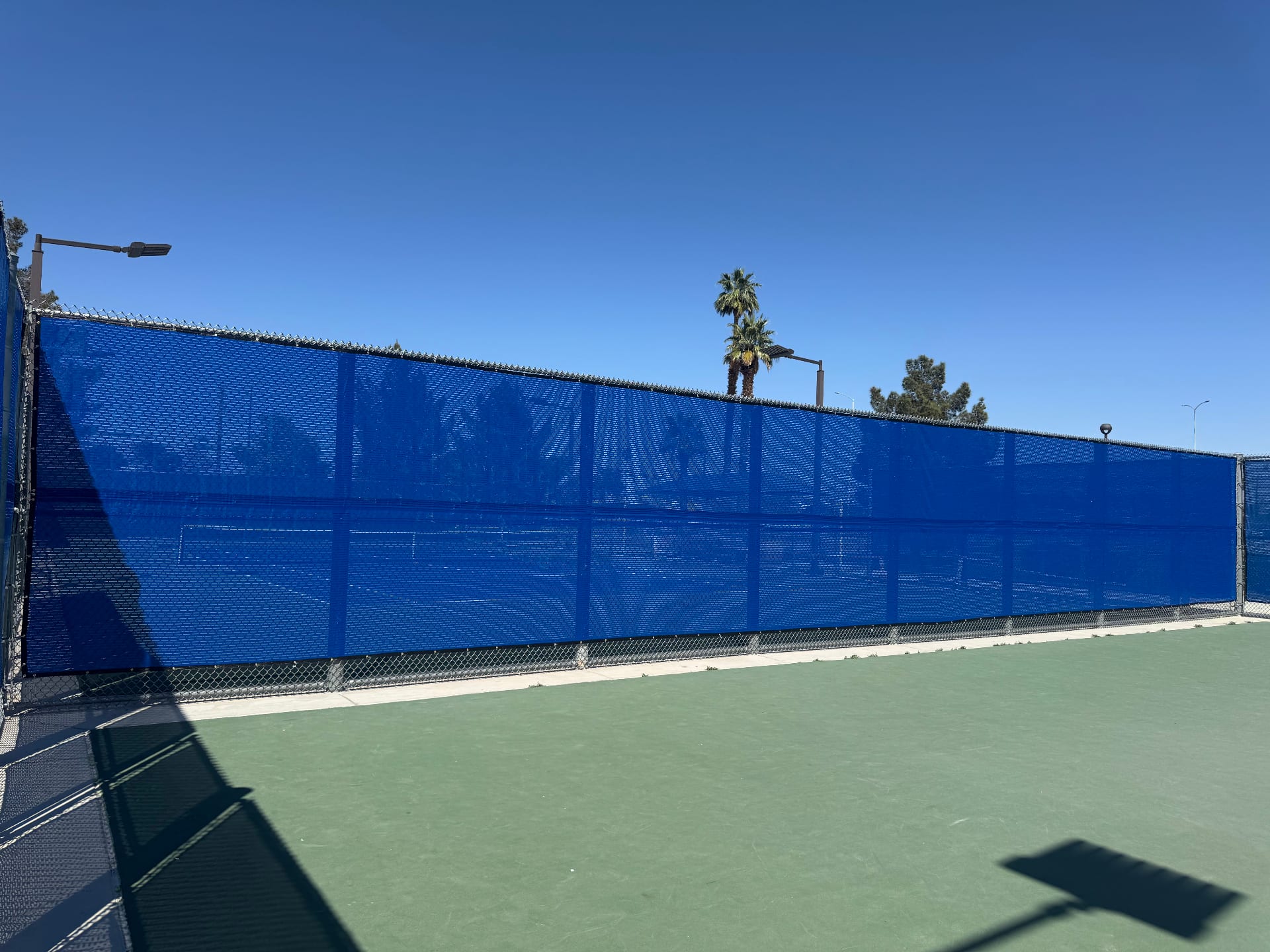 Bright blue custom windscreen installed on a chain link fence surrounding a green athletic court in Henderson, Nevada.