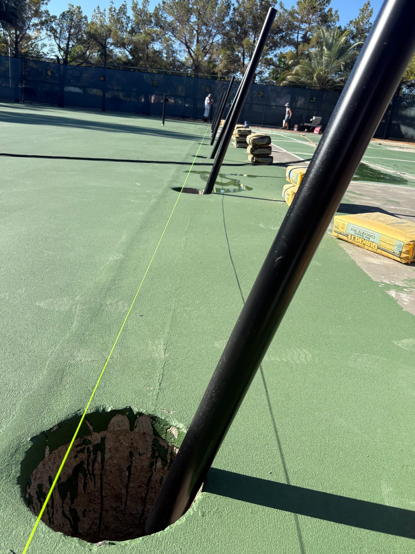 Installation of new black metal fence posts on a green athletic court in Las Vegas with concrete mix bags visible.