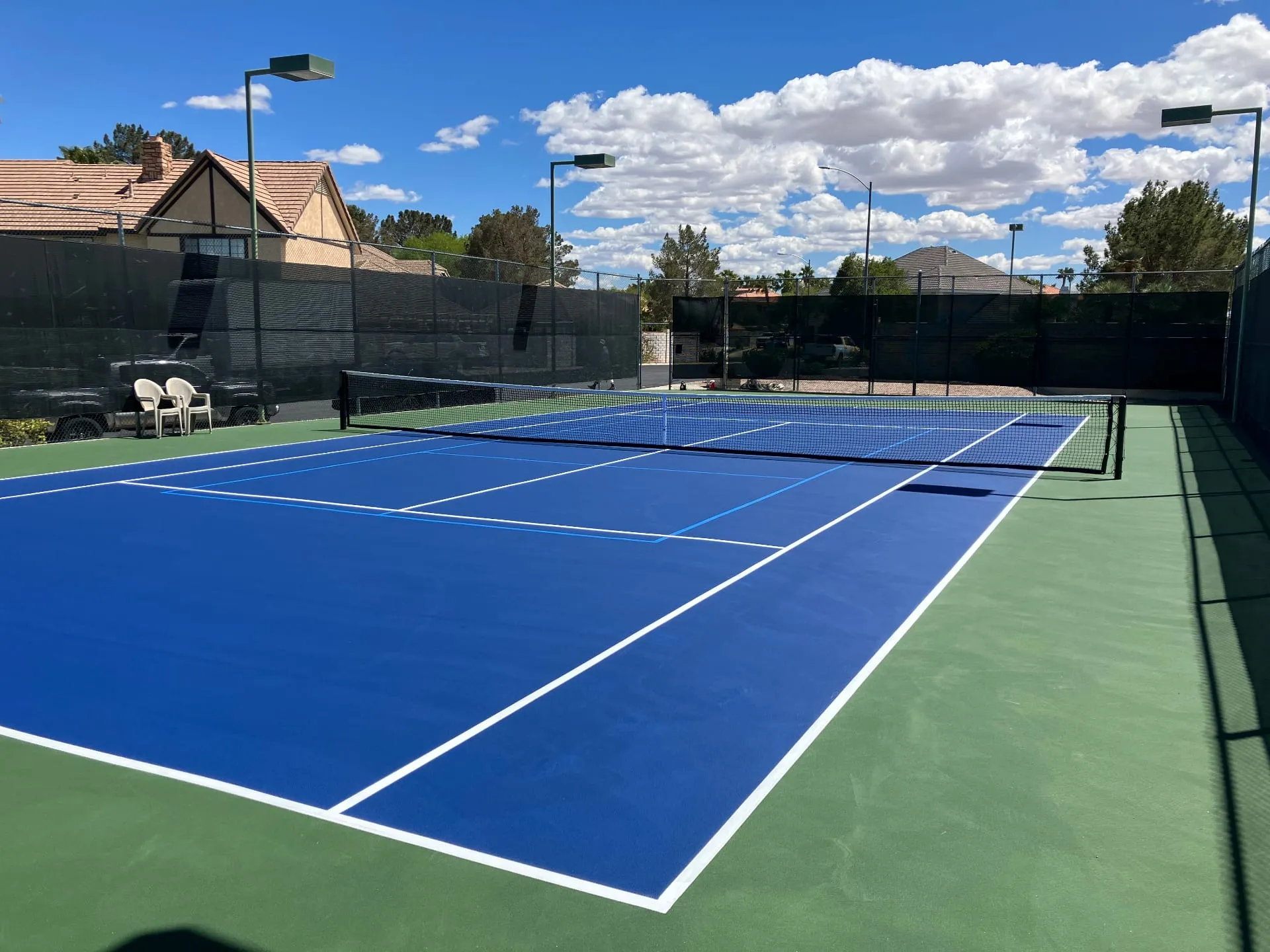 A freshly resurfaced blue and green tennis court in a residential backyard with black windscreen and court lighting.